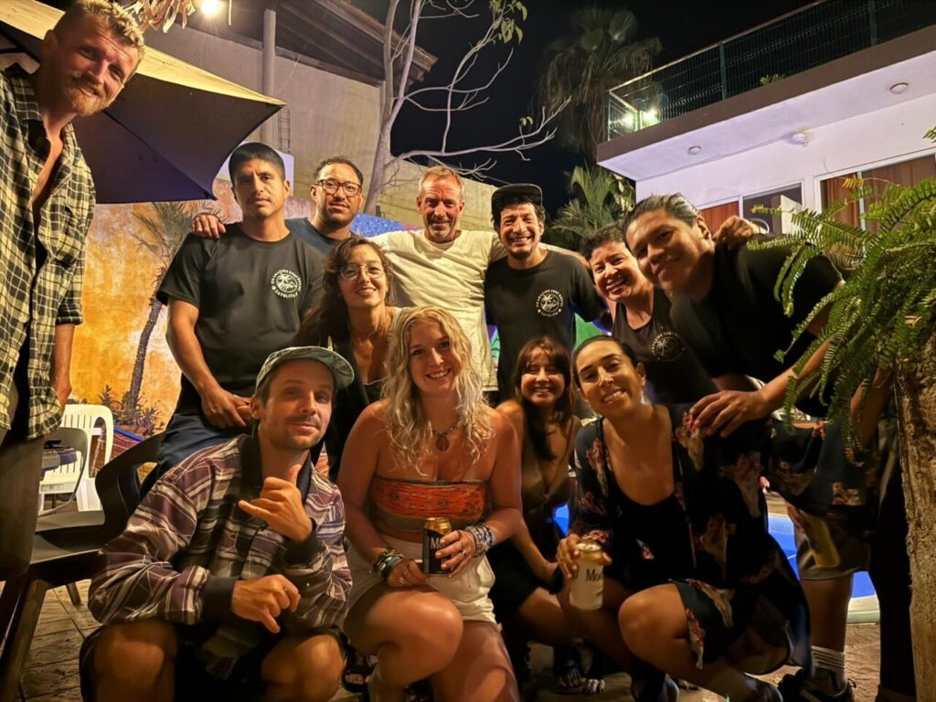 A diverse group of international travelers and staff smile and pose closely together during an evening outdoor social gathering at a tropical backpacker hostel.