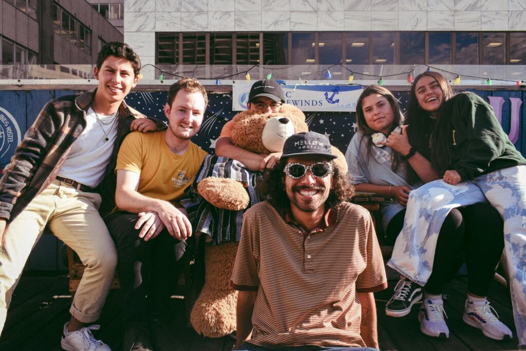 A diverse group of young international backpackers smiling and posing together on an outdoor hostel rooftop patio decorated with string lights, featuring a large teddy bear.