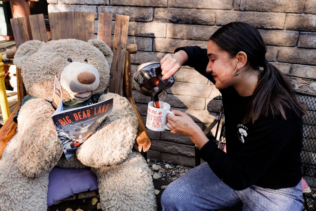 A solo traveler pouring coffee next to a giant teddy bear reading a Big Bear Lake guidebook on a patio chair at ITH Big Bear Mountain Eco Lodge