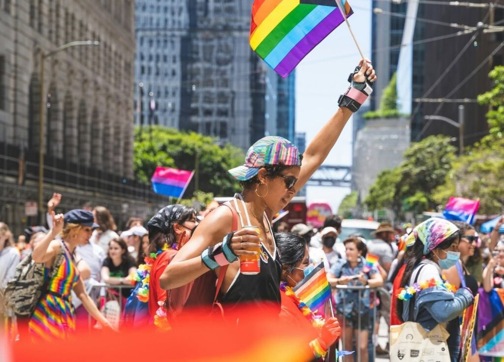 A person celebrates at the San Francisco Pride Parade in June, waving a rainbow pride flag high in the air while wearing wrist guards, a colorful cap, and holding a drink amid a crowded street festival.