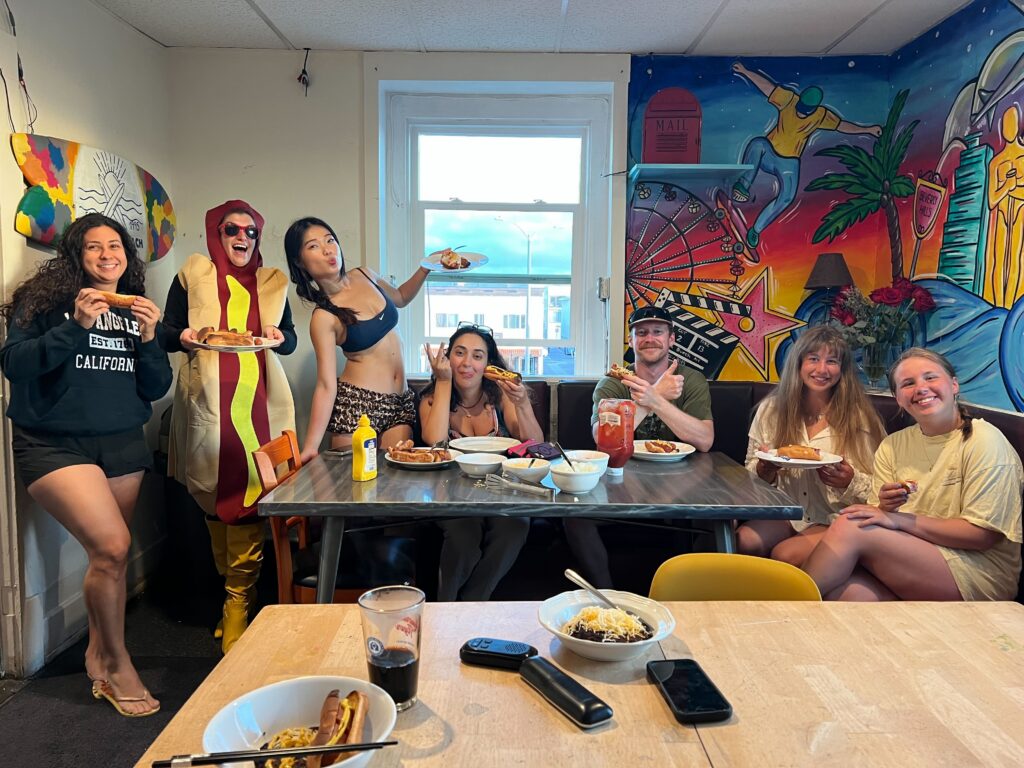 A diverse group of young backpackers smiles and poses around a dining table inside a hostel common room, enjoying a hot dog meal with one person playfully dressed in a full hot dog costume next to a colorful mural wall.