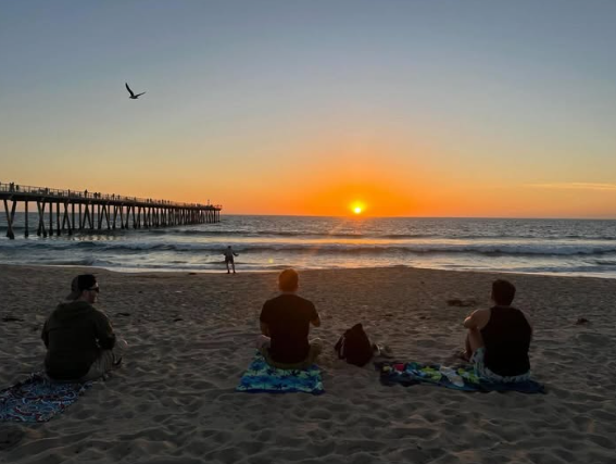 Three backpackers sit on towels on the sandy beach watching a vibrant orange sunset over the ocean next to the Hermosa Beach Pier.